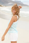 A young blonde woman on a beach wearing a light-blue top and white summer shorts