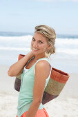 A mature blonde woman on a beach wearing a white top and a turquoise vest with a wicker basket