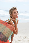 A mature blonde woman on a beach wearing a white top and a turquoise vest with a wicker basket