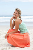 A mature blonde woman on a beach wearing a white top, a turquoise vest and a salmon coloured skirt