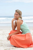 A mature blonde woman on a beach wearing a white top, a turquoise vest and a salmon coloured skirt