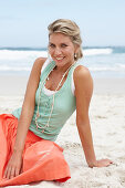 A mature blonde woman on a beach wearing a white top, a turquoise vest and a salmon coloured skirt