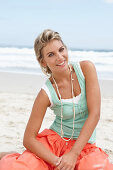 Reife blonde Frau im weißen Top, türkisfarbener Weste und lachsfarbenem Rock am Strand