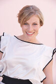 A blonde woman standing against a pink background wearing a light blouse with a black trim