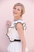 A blonde woman standing against a pink background wearing a light blouse with a black trim