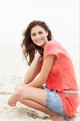 A brunette woman on a beach wearing a salmon-coloured top and a denim skirt