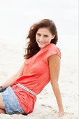 A brunette woman on a beach wearing a salmon-coloured top and a denim skirt