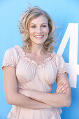 A blonde woman standing against a blue surface wearing a pink short-sleeved blouse