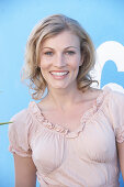 A blonde woman standing against a blue surface wearing a pink short-sleeved blouse