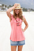 A young blonde woman on a beach wearing a pink top, a short denim skirt and a beige hat