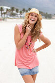 Junge blonde Frau im rosa Top, kurzem Jeansrock und beigem Hut am Strand