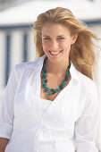 A young blonde woman on a beach wearing a white blouse and a chunky necklace