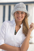 A young blonde woman on a beach wearing a white blouse and a white crocheted hat