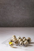 Raw uncooked quail eggs whole and broken in plastic boxing on white marble kitchen table