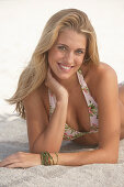 Junge blonde Frau im gemusterten Bikini liegt am Sandstrand
