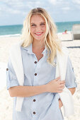 A young blonde woman on a beach wearing a striped shirt with a white jumper over her shoulders