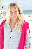A young blonde woman on a beach wearing a striped shirt with a pink jumper over her shoulders