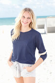 A young blonde woman on a beach wearing a long-sleeved blue shirt and shorts