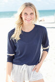 A young blonde woman on a beach wearing a long-sleeved blue shirt and shorts