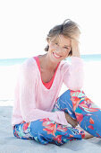 A mature woman with short blonde hair on a beach wearing a pink and white top and colourful trousers