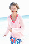 A mature woman with short blonde hair on a beach wearing a pink and white top and colourful trousers