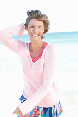 A mature woman with short blonde hair on a beach wearing a pink and white top and colourful trousers