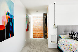 Bett mit bunter Bettwäsche, Nachtkästchen und Pendelleuchte, an Wand moderne Malerei im Schlafzimmer mit Durchgang