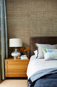Double bed with leather headboard, bedside table with lamp and textile wallpaper in the bedroom
