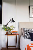 Bedside table with lamp, branch of leaves and books next to bed with headboard