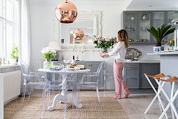 Open-plan kitchen with pale grey cupboards, round dining table and designer chairs - woman holding vase of roses