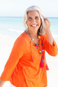 A mature woman with white hair on a beach wearing an orange tunic