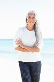 A mature woman with white hair on a beach wearing a white jumper and polka dot trousers