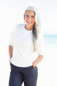 A mature woman with white hair on a beach wearing a white jumper and polka dot trousers