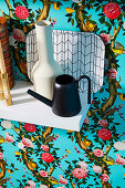 Vase, small watering can and tray on white wall shelf in front of floral wallpaper