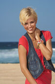 A blonde woman with short hair by the sea wearing a red top and a denim gilet
