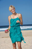 A blonde woman with short hair on a beach holding a pitahaya wearing a turquoise dress