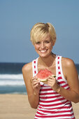 A blonde woman with short hair by the sea wearing a red-and-white striped top and holding a slice of watermelon