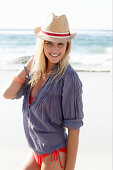 A blonde woman on a beach wearing a hat, a red bikini and a blue blouse