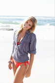 A blonde woman on a beach wearing a red bikini and a blue blouse