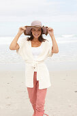 A brunette woman by the sea wearing a short-sleeved cardigan and a hat