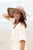 A brunette woman by the sea wearing a short-sleeved cardigan and a hat