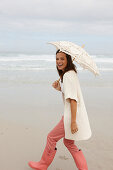 Brünette Frau mit Sonnenschirm in Strickjacke, rosa Hose und Gummistiefeln am Meer