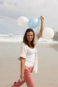 Brünette Frau mit Luftballons in Strickjacke und rosa Hose am Meer