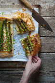 A hand taking a piece of puff pastry tart with green asparagus and onions