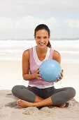 A young brunette woman by the sea with a ball sitting in the lotus position