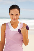 A young brunette woman by the sea wearing sports clothes with a dumbbell