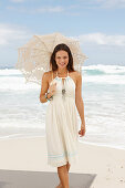 A brunette woman by the sea wearing a white summer dress and holding a parasol