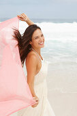 A brunette woman by the sea wearing a white summer dress and holding a pink shawl