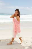 A brunette woman by the sea wearing a white summer dress and holding a pink shawl