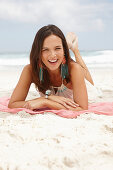 A brunette woman lying on a pink shawl by the sea wearing a white summer dress